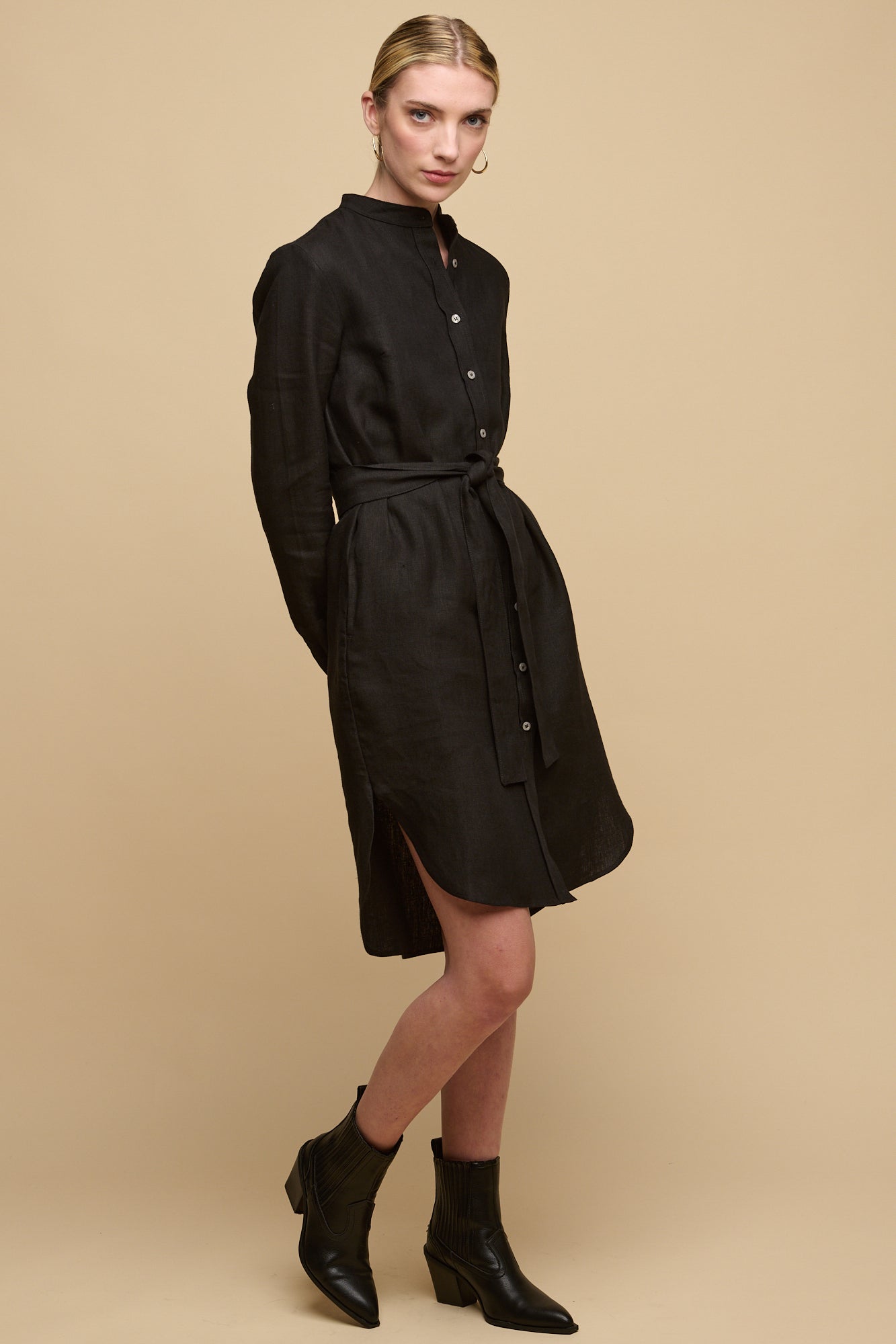 Full body image of collarless shirt dress in black linen with waist belt tied and paired with black boots