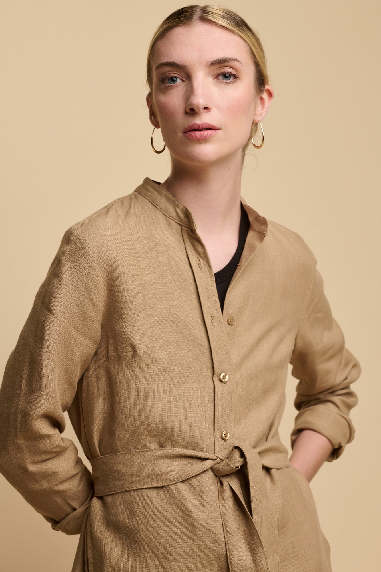 Hip up image of female wearing collarless shirt dress in truffle linen with belt tied at waist and sleeves rolled up slightly. worn over black vest