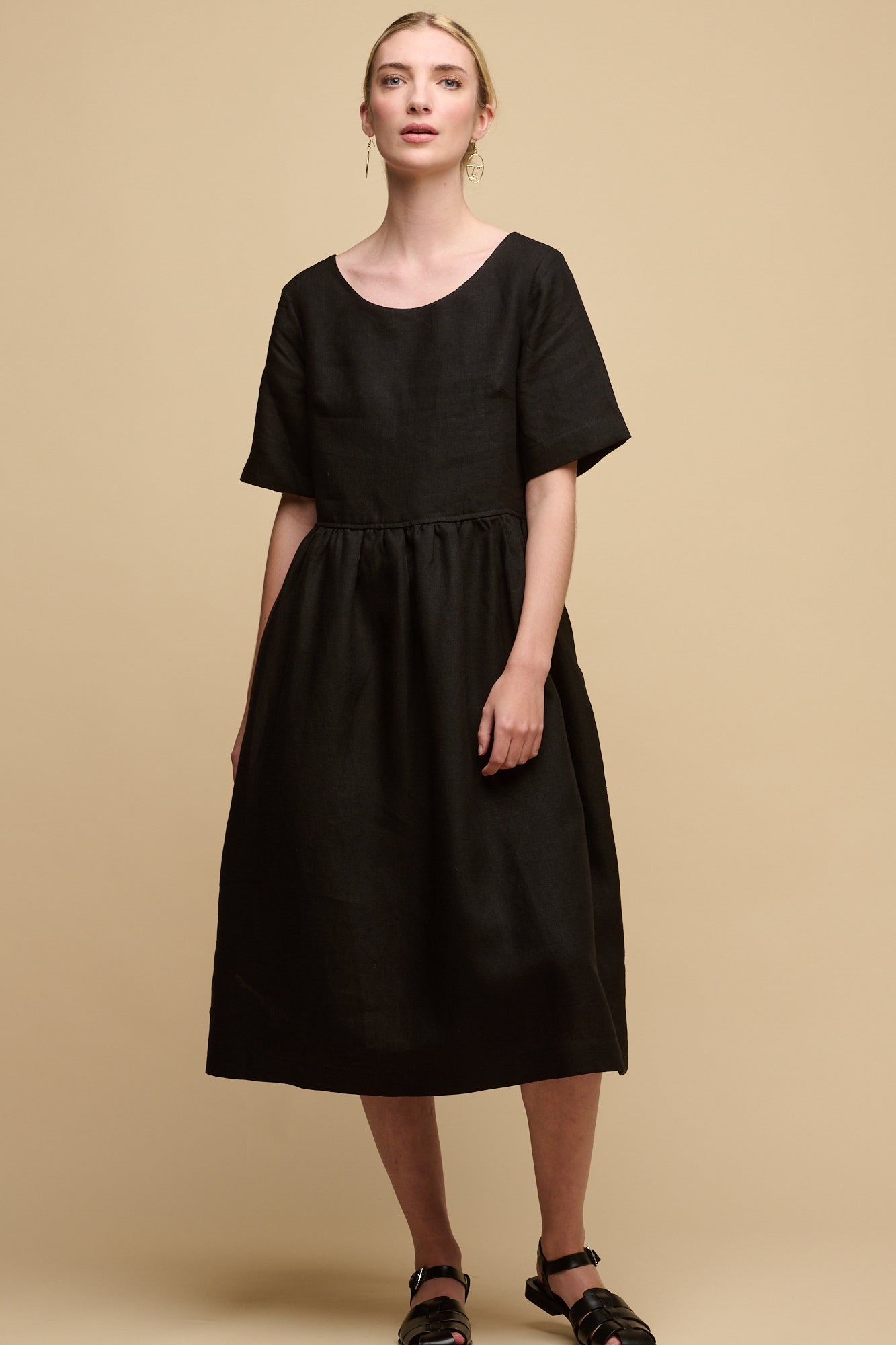 Full body image of fair skinned female with hair tied back into a bun wearing crew neck gathered dress in black linen