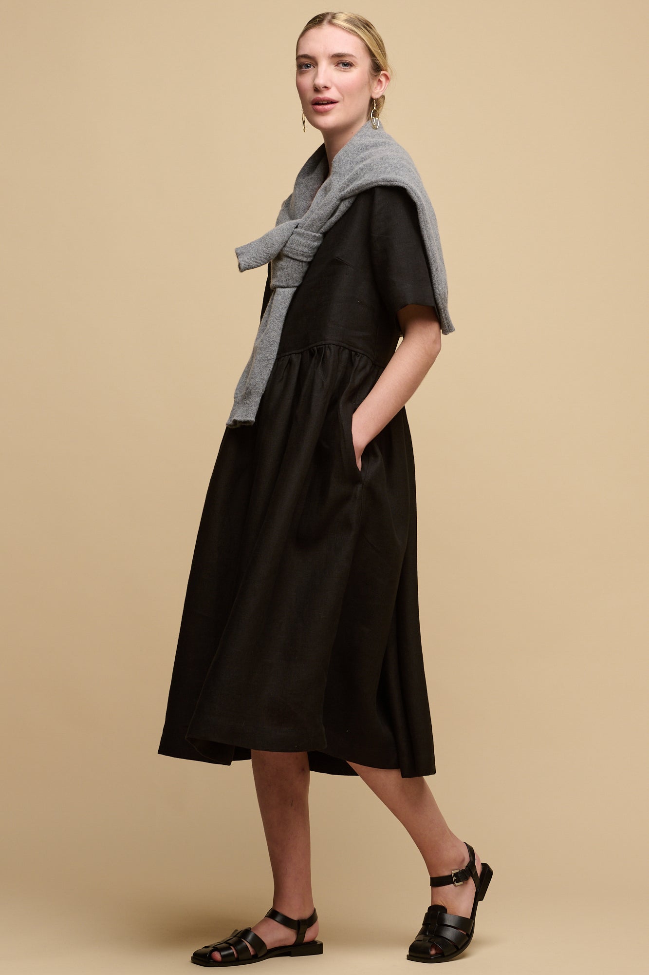 Full body image of fair skinned female with hair tied back into a bun wearing crew neck gathered dress in black linen with lambswool jumper draped over the shoulders and paired with sandels.