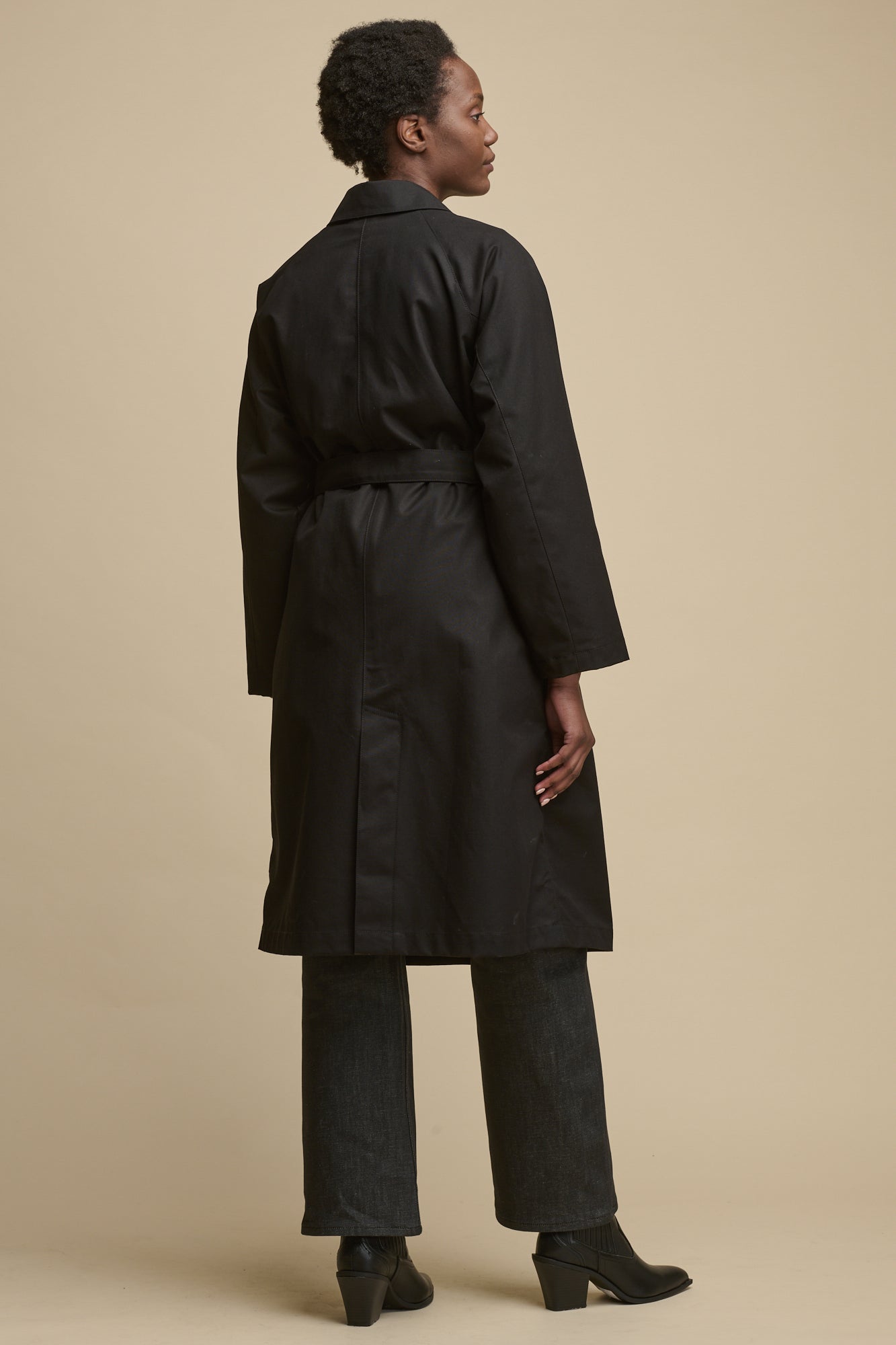 Full body image of the back of female wearing women's Frances Raglan Belted Raincoat in black paired with black jeans and black boots