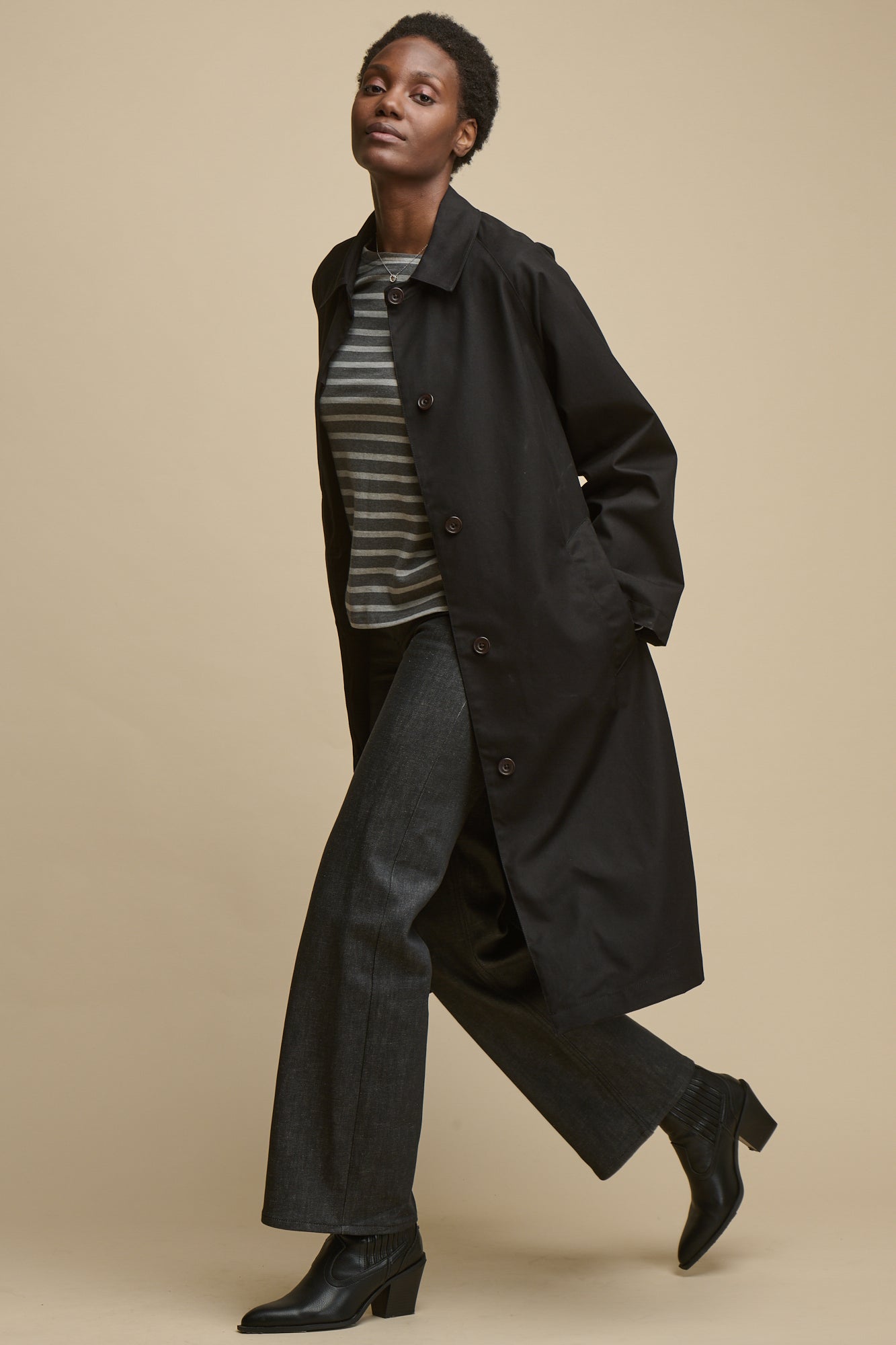 Full body image of female walking wearing women's Frances Raglan Belted Raincoat in black unfastened worn with Breton in charcoal marl and grey marl