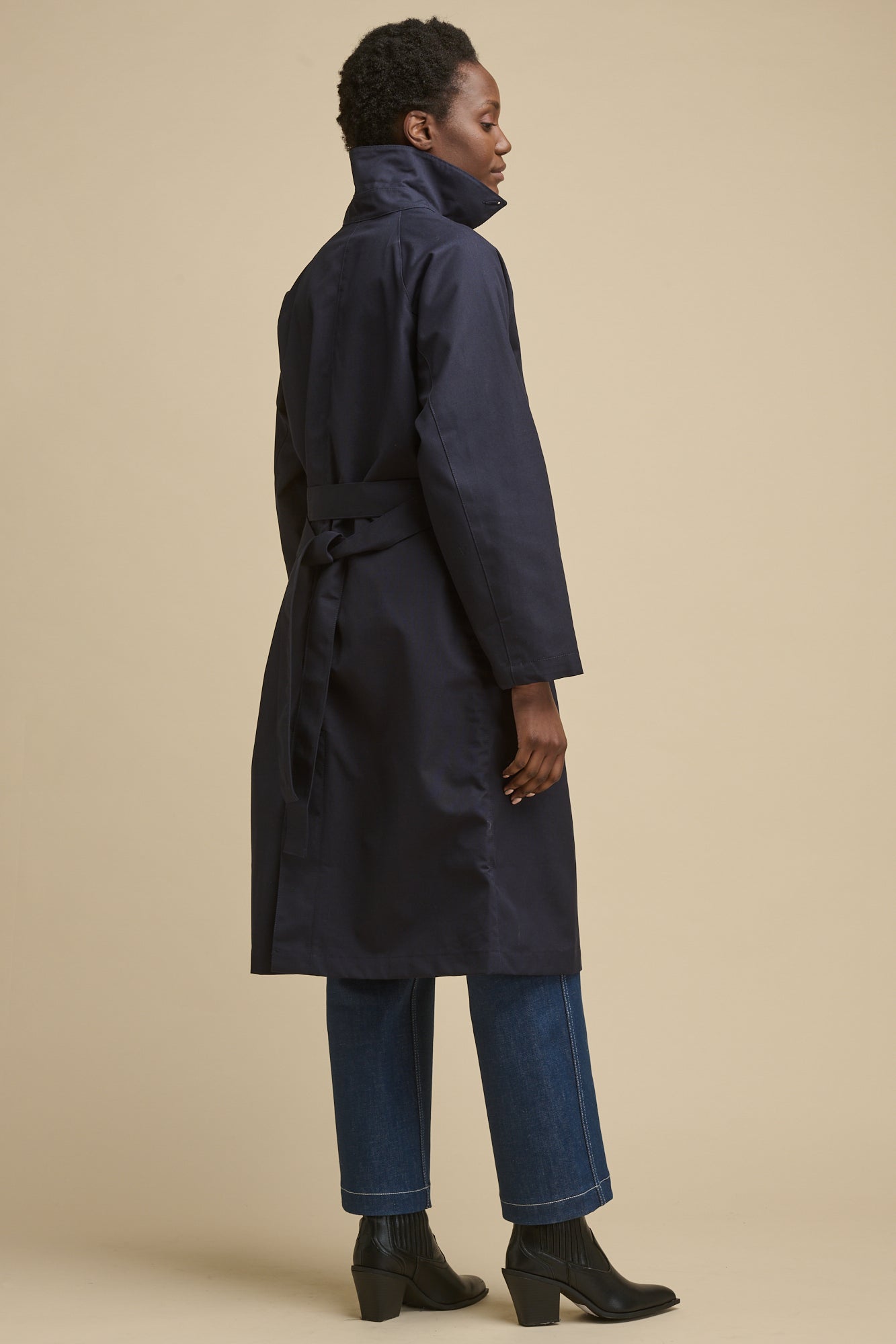 Full body image of the back of female wearing women's Frances Raglan Belted Raincoat in navy with belt tied at the back and collar up