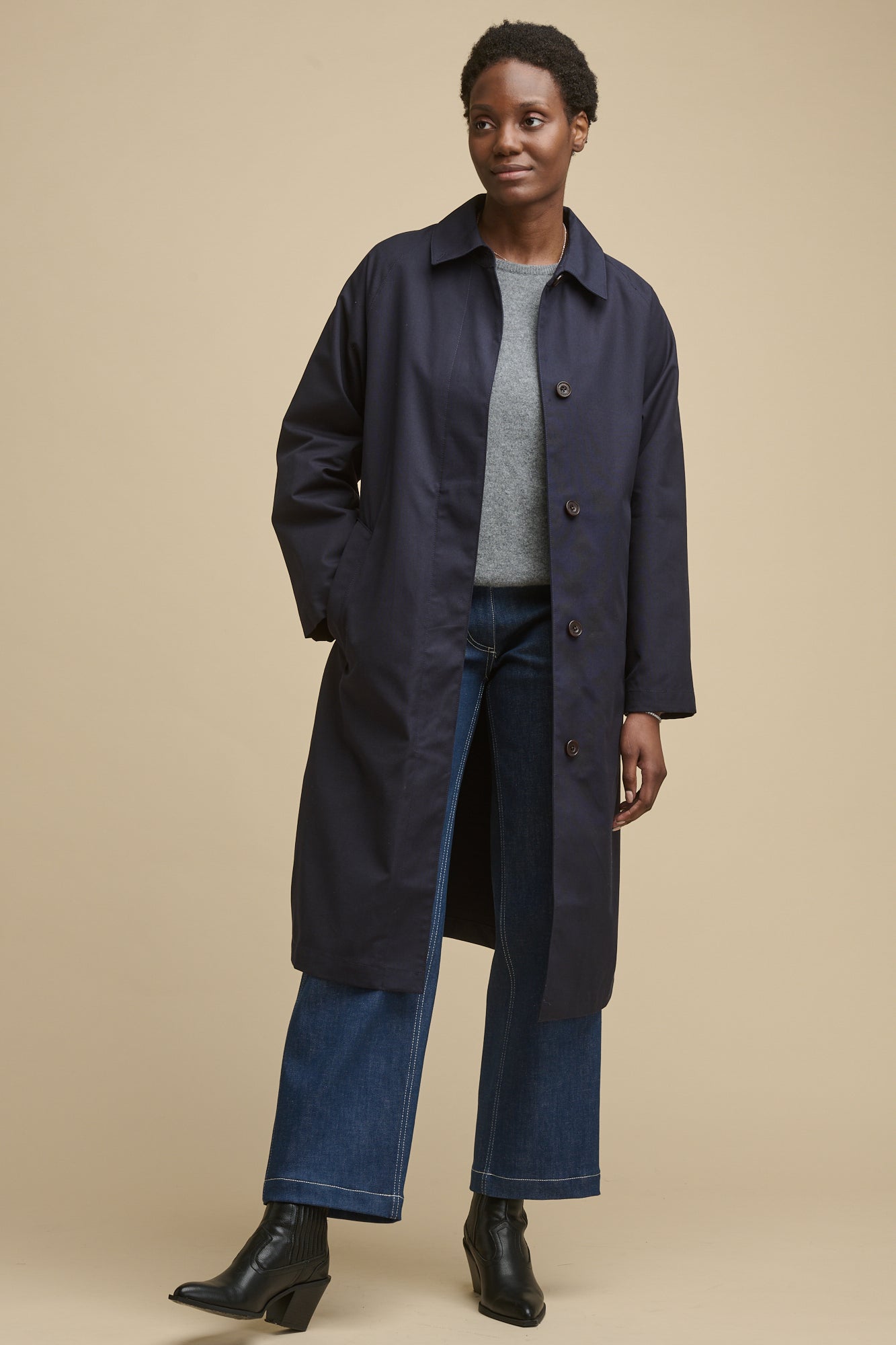 Full body image of female wearing women's Frances Raglan Belted Raincoat in navy unfastened over grey lamsbwool crew neck jumper and blue jeans