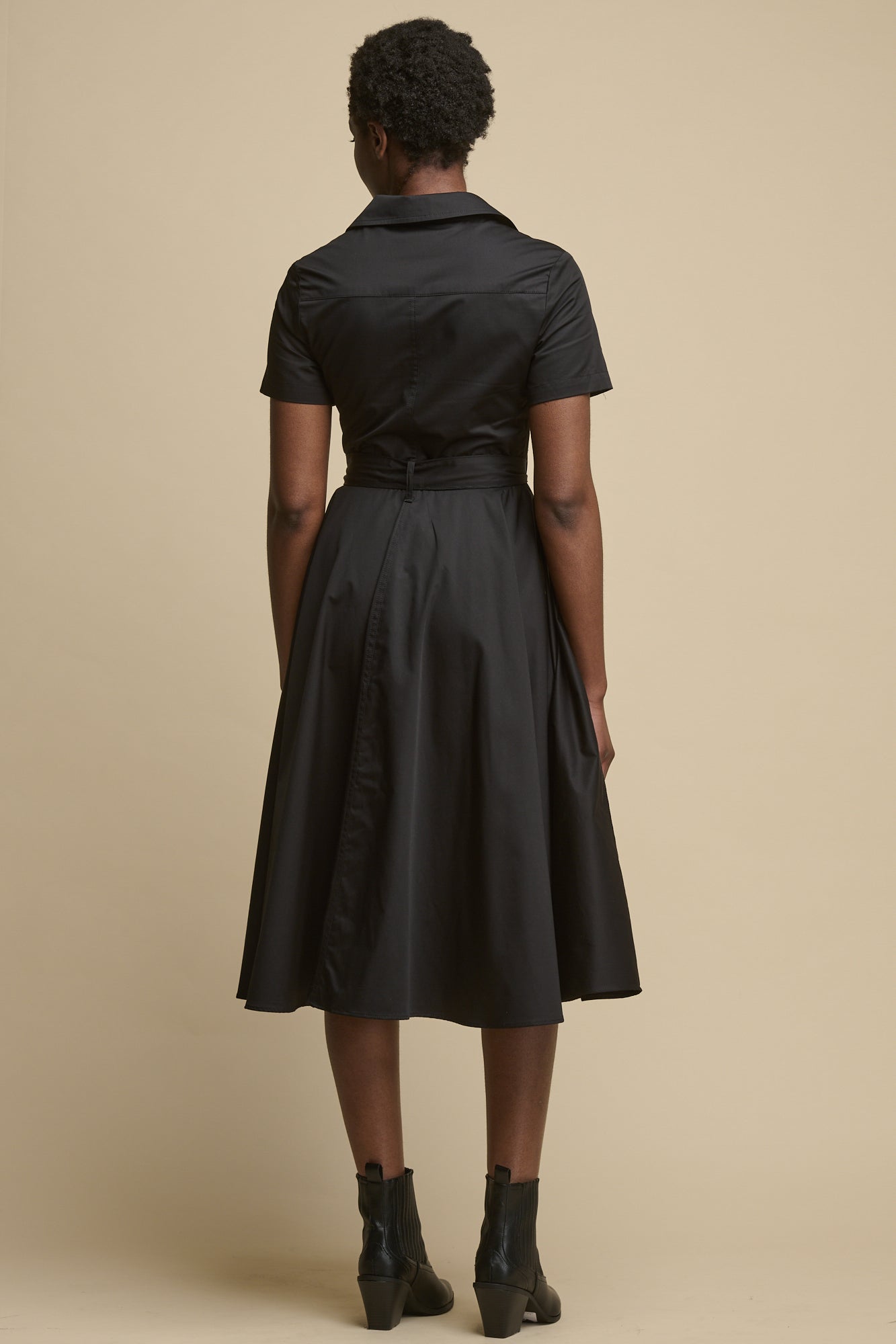 Full body image of the back of black female wearing full skirted phoebe trench dress in black, paired with black boots