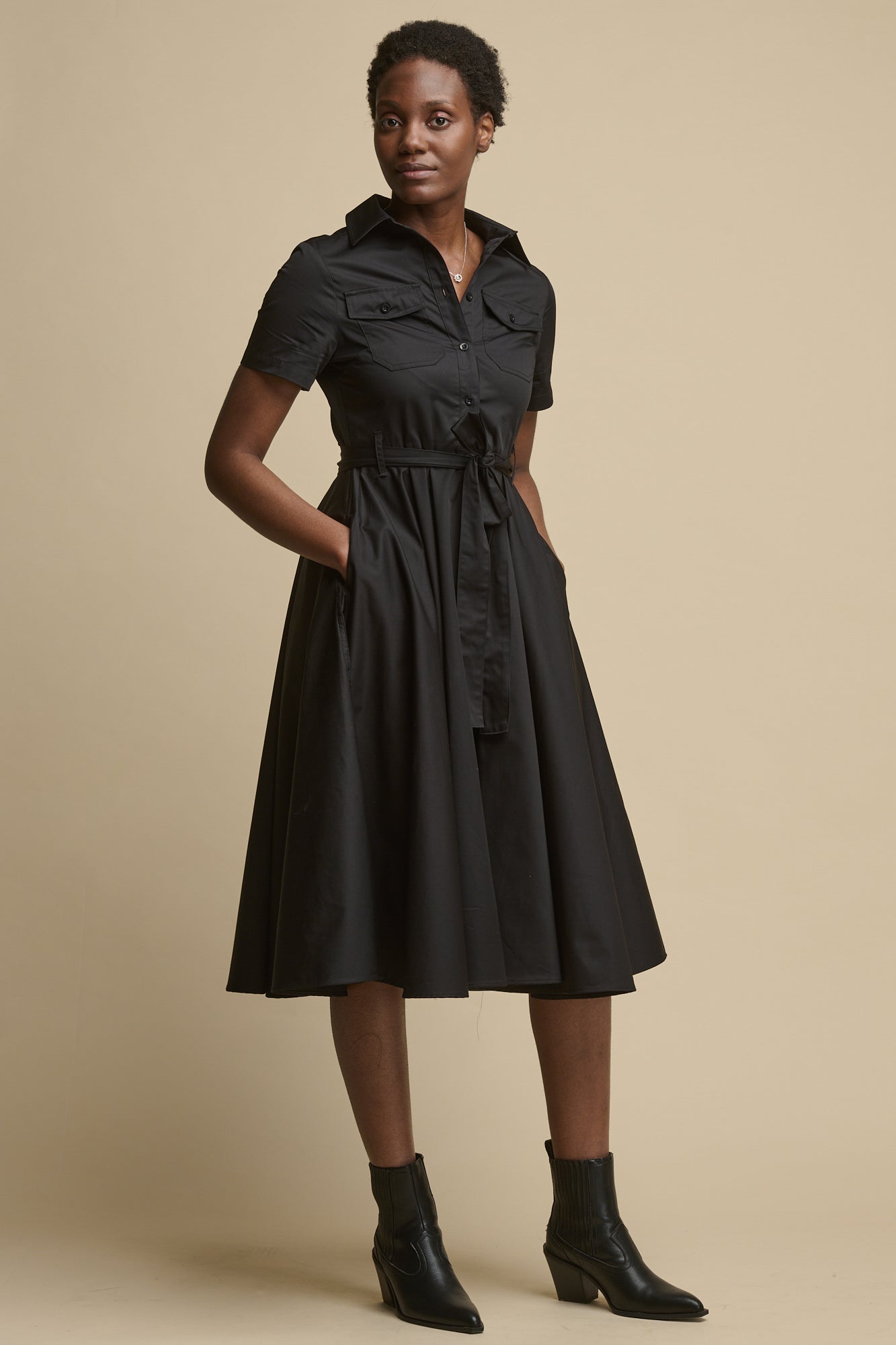 Full body image of black female wearing full skirted phoebe trench dress in black, with the belt tied in a bow and her hands in the pockets, worn with black boots