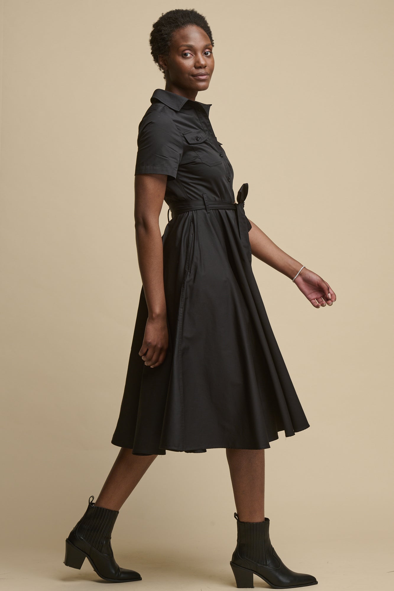 Full body image of the side of female mid walking stance looking to the camera wearing full skirted phoebe trench dress in black with short sleeves and belt tied into a bow 