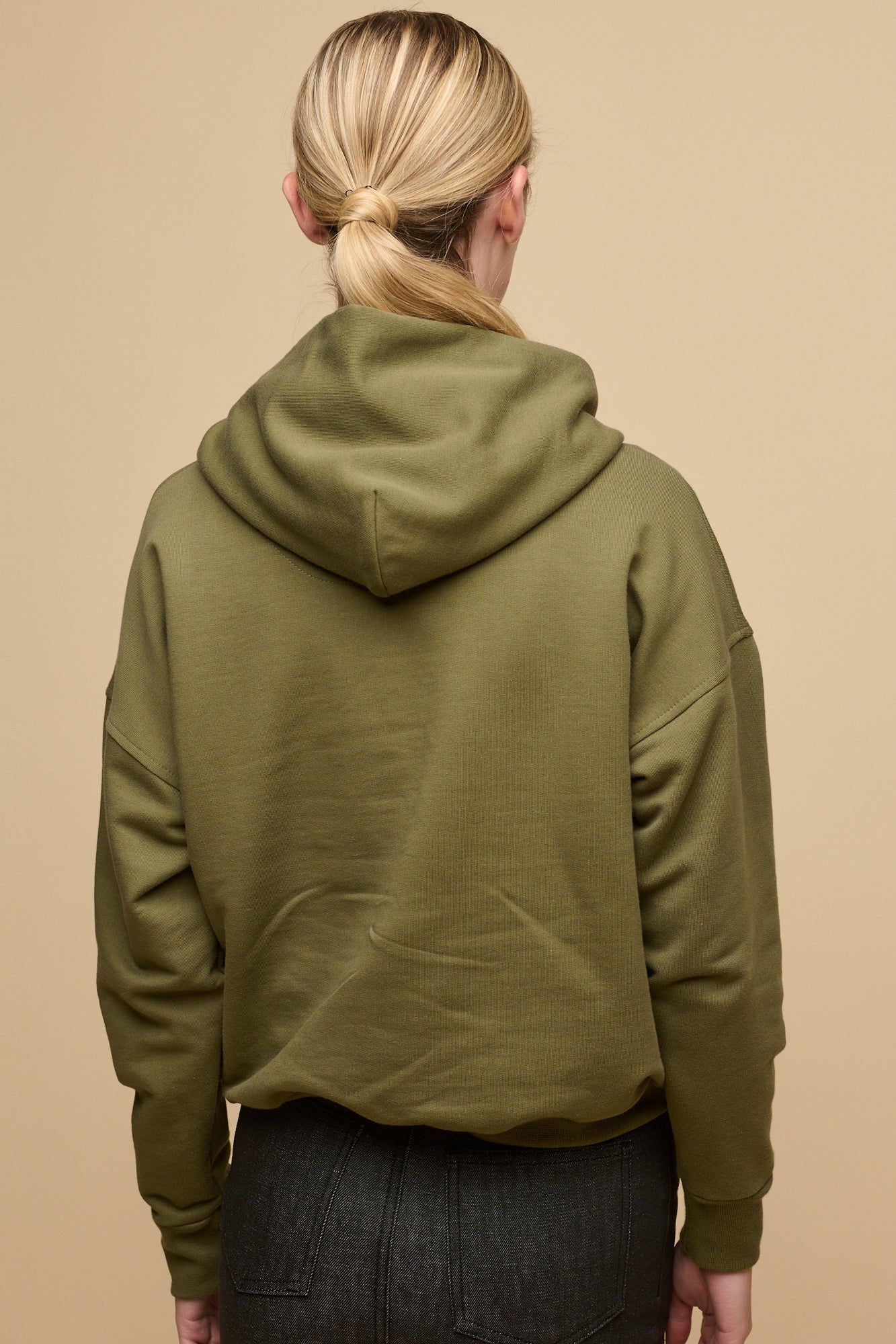 The back of female with blonde hair tied back into a low ponytail wearing hooded sweatshirt in olive 