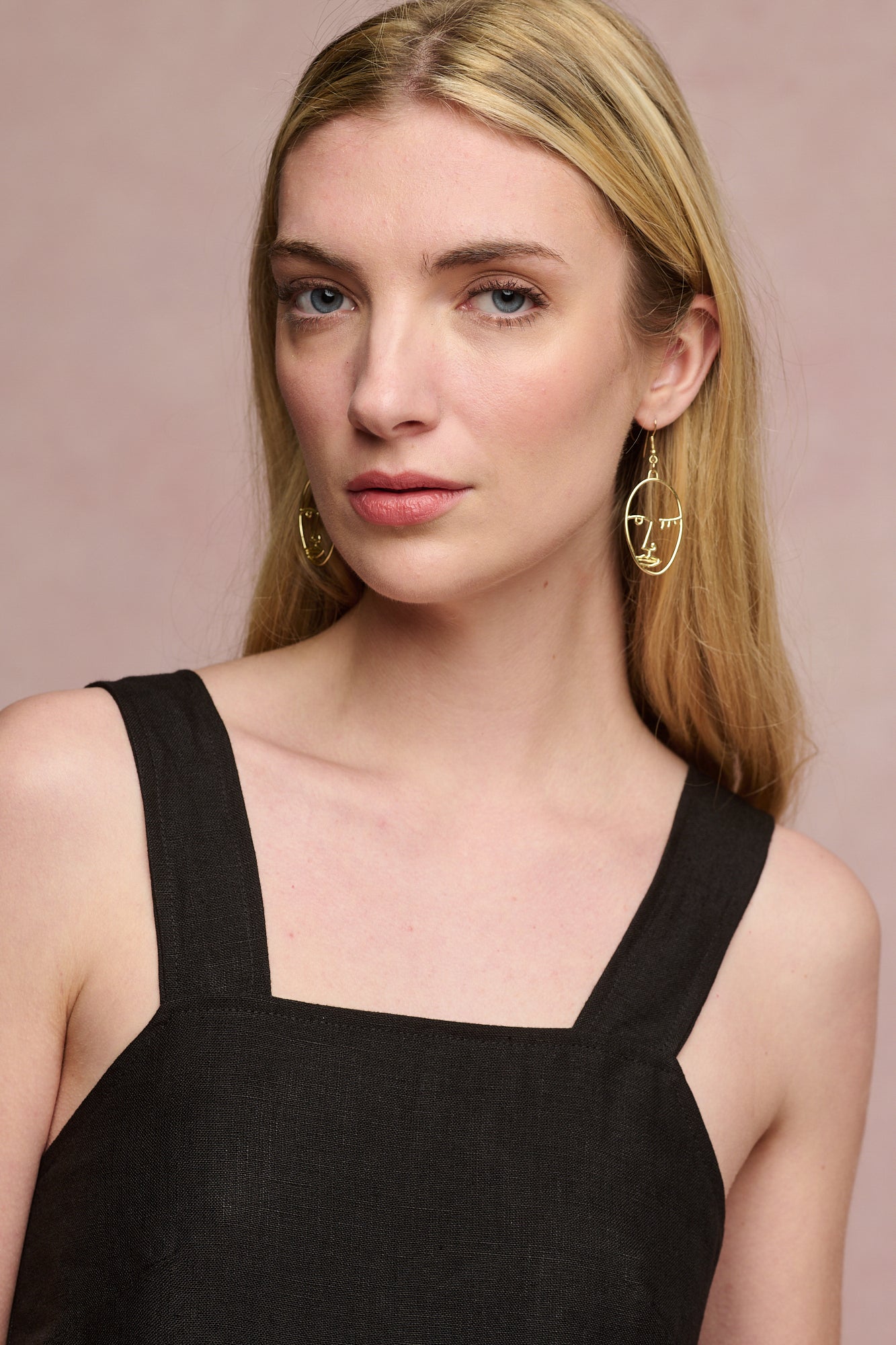 Portrait of fair skinned female with blonde hair behind ears wearing linen sun dress in black with gold earrings.