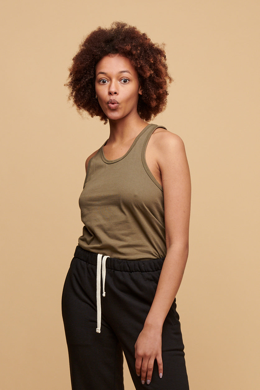 Female from the thigh up wearing racer back vest in olive made form 100% cotton  tucked into black sweatpants also made from 100% cotton