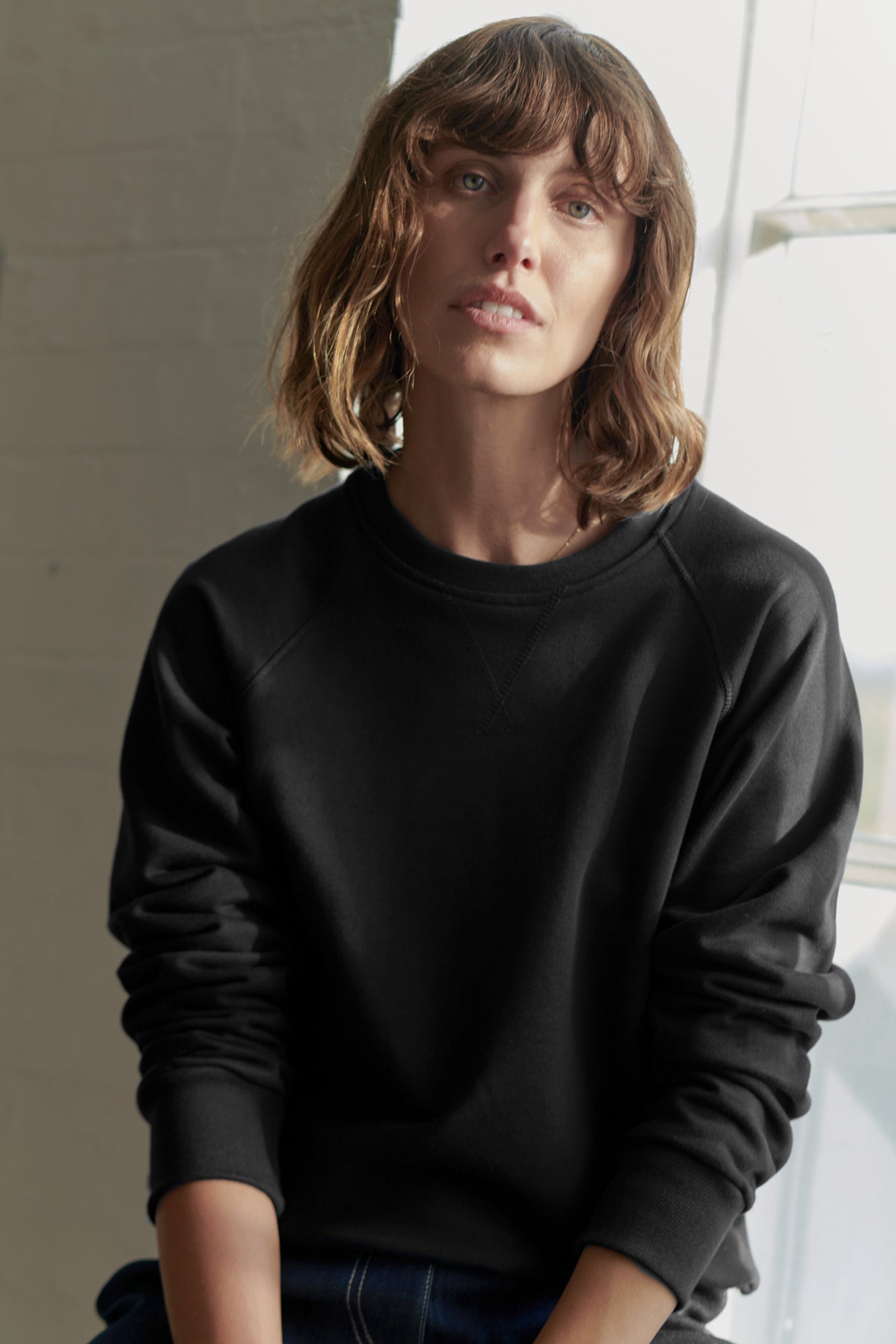 Thigh up image of brunette, white female wearing women's raglan sweatshirt in black with the sleeves pushed up